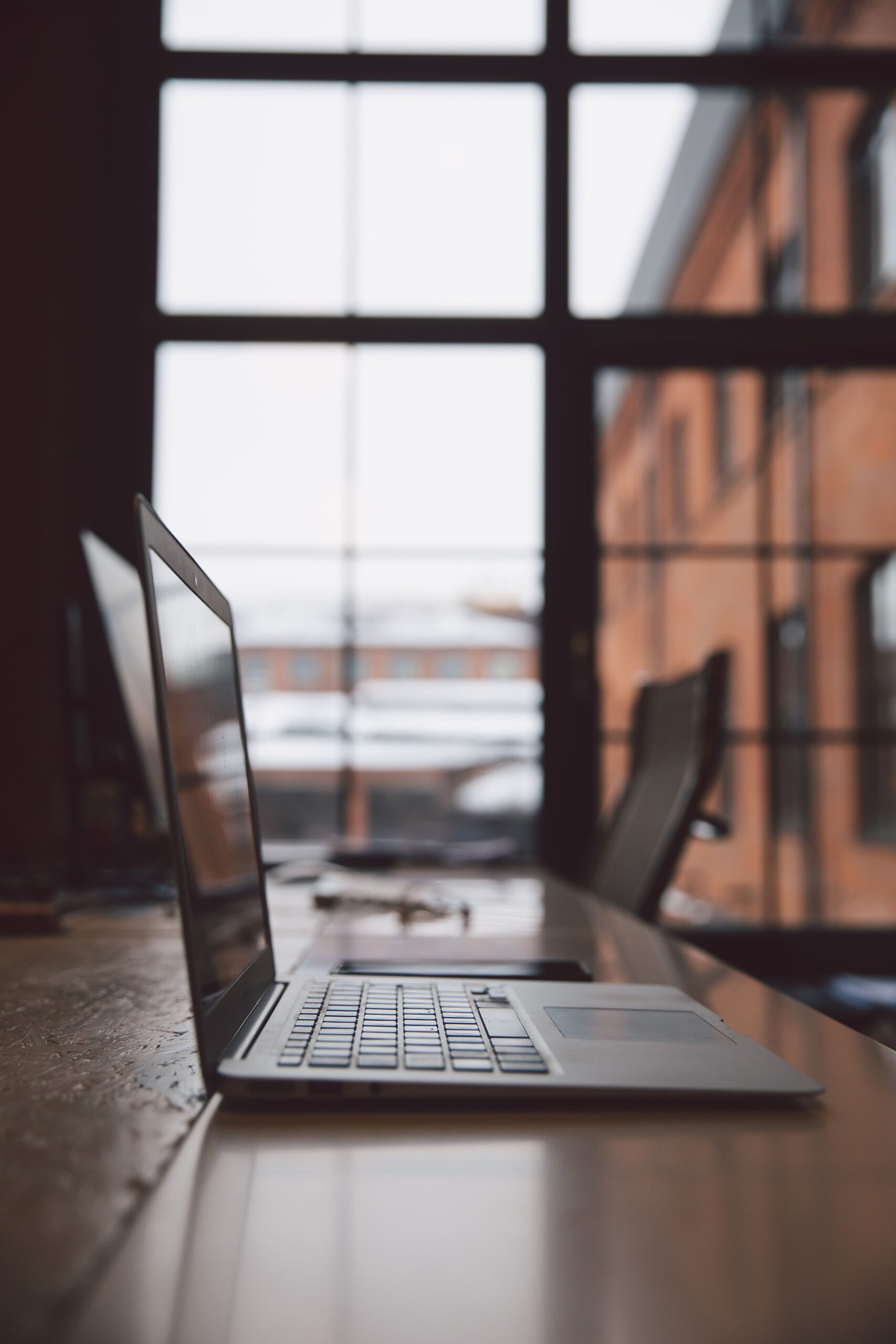 Minimalist office with a laptop on a wooden desk in front of large windows providing natural light.
